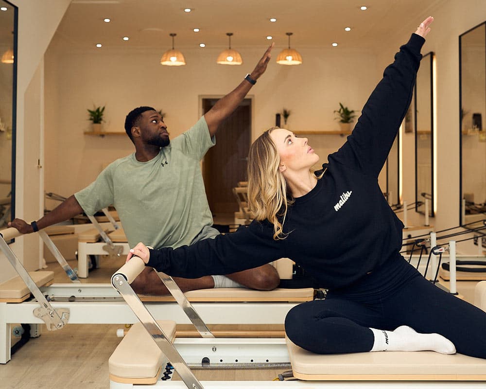 Two members seated in a side reach on the reformers at Malibu Studios, Oldswinford